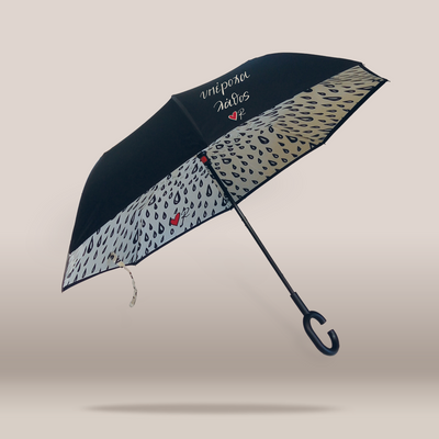 A floating umbrella with a black exterior and a light beige interior patterned with dark raindrops. The inside canopy features white script text reading 'Interacl Taxis' with a small red heart, and the handle is a dark hook-style design.