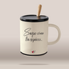 An off-white ceramic mug with a black lid and handle, featuring black Greek script text and a small red heart logo near the bottom. A light wood spoon rests inside the mug, which is set against a solid beige background.
