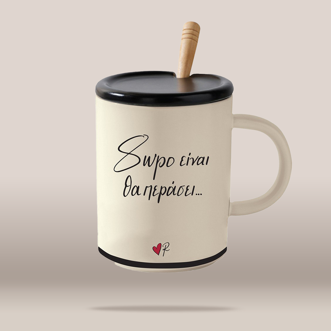 An off-white ceramic mug with a black lid and handle, featuring black Greek script text and a small red heart logo near the bottom. A light wood spoon rests inside the mug, which is set against a solid beige background.