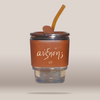 A clear glass tumbler with a brown faux-leather sleeve printed with white cursive text, topped with a brown lid and a translucent brown straw, shown on a plain beige background.