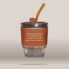A clear glass tumbler with a brown leather-like sleeve and a translucent brown straw, shown floating on a plain warm beige background. White Greek text is printed on the sleeve.