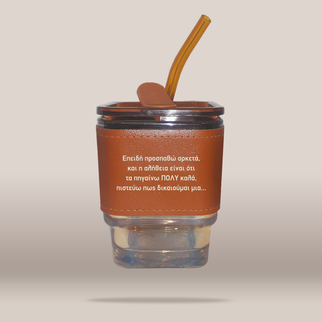A clear glass tumbler with a brown leather-like sleeve and a translucent brown straw, shown floating on a plain warm beige background. White Greek text is printed on the sleeve.