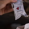 Hand holding a small white packet with a red heart and letter 'P' logo, against a blurred background.