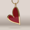 A heart-shaped keychain with a glossy red enamel center and a polished gold-tone metal border, attached to a gold split ring, shown against a neutral beige background.
