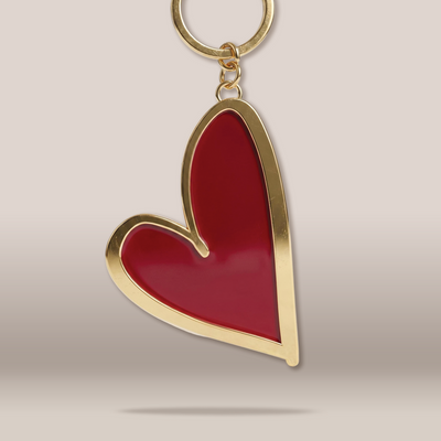 A heart-shaped keychain with a glossy red enamel center and a polished gold-tone metal border, attached to a gold split ring, shown against a neutral beige background.