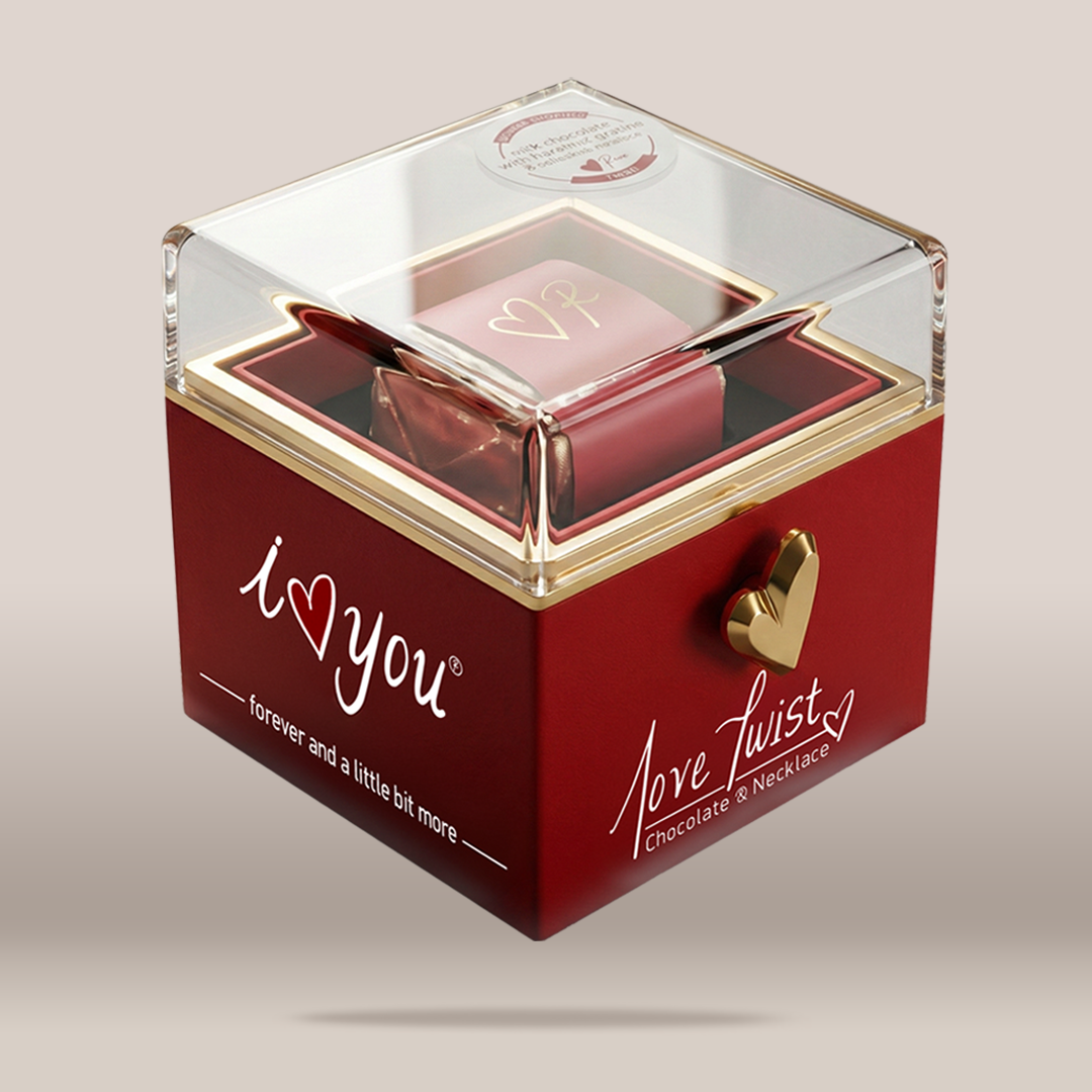 A red square chocolate gift box with a clear acrylic lid, gold trim, and a gold heart-shaped clasp. The front of the box reads 'i <heart> you' and 'Love Twist Chocolate & Necklace,' with a small necklace visible inside.
