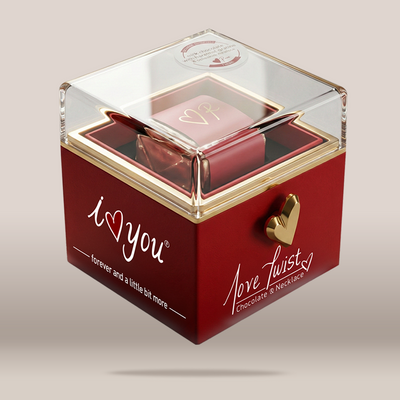 A red square chocolate gift box with a clear acrylic lid, gold trim, and a gold heart-shaped clasp. The front of the box reads 'i <heart> you' and 'Love Twist Chocolate & Necklace,' with a small necklace visible inside.