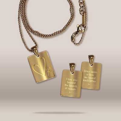 Three gold-toned pendant necklaces arranged on a light beige studio background. One larger rectangular pendant on a beaded chain shows an engraved heart and initials, while two smaller matching pendants with short chains display engraved romantic phrases.