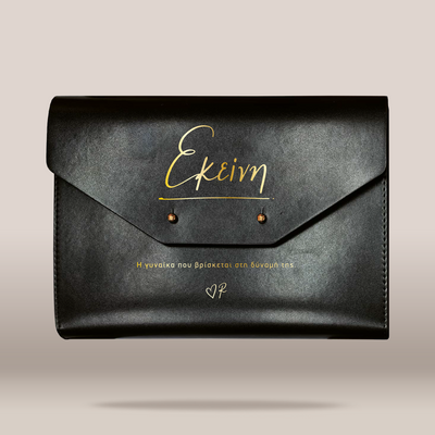 A black leather envelope-style clutch bag with gold foil script lettering on the front flap, photographed against a plain light beige background.