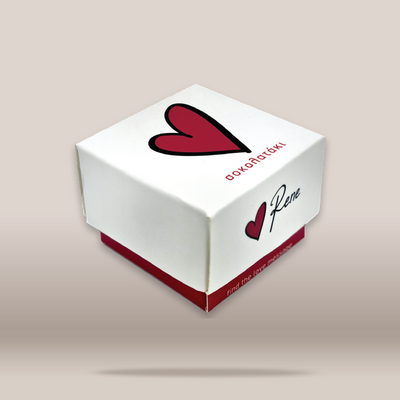 A white square gift box with a red heart logo and the text 'OkoŁătki' and 'Paweł' on the lid, shown floating against a light beige background.