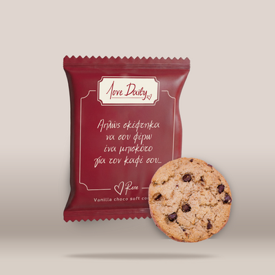 A red stand-up pouch with Greek text and the brand name 'Love Daily' stands next to a round chocolate chip cookie on a light beige background.