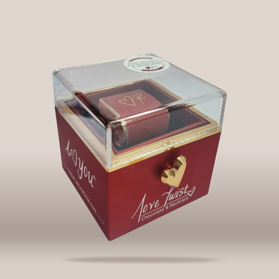 A square-shaped red chocolate gift box with a clear plastic top lid, featuring a gold heart-shaped clasp and white script text that reads 'Love Twisty Chocolate & Treats' on the front, set against a plain beige background.