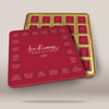 A red square gift box with a hinged lid, shown in front of an open gold-toned tray containing multiple small red cards or inserts. The lid has white script text reading 'Love Reasons' and Greek text arranged around the perimeter.