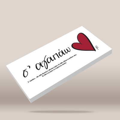 A white rectangular card or box shown at an angle on a warm beige background. The front has black cursive Greek text and a large red heart graphic.