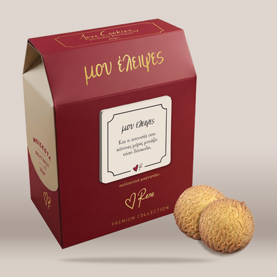A red gift box of Greek cookies with gold lettering and a white label, shown next to two round golden-brown cookies on a light beige background.