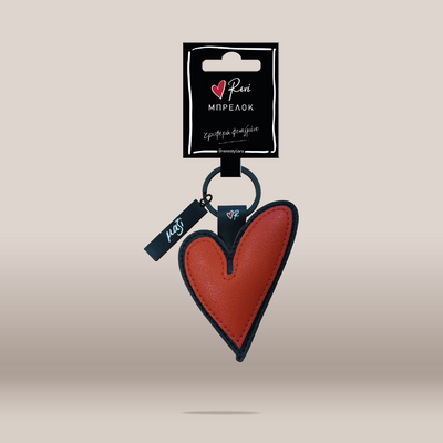 A red leather heart-shaped keychain with black trim, attached to a black key ring and a small black tag, hanging from a black retail card. 