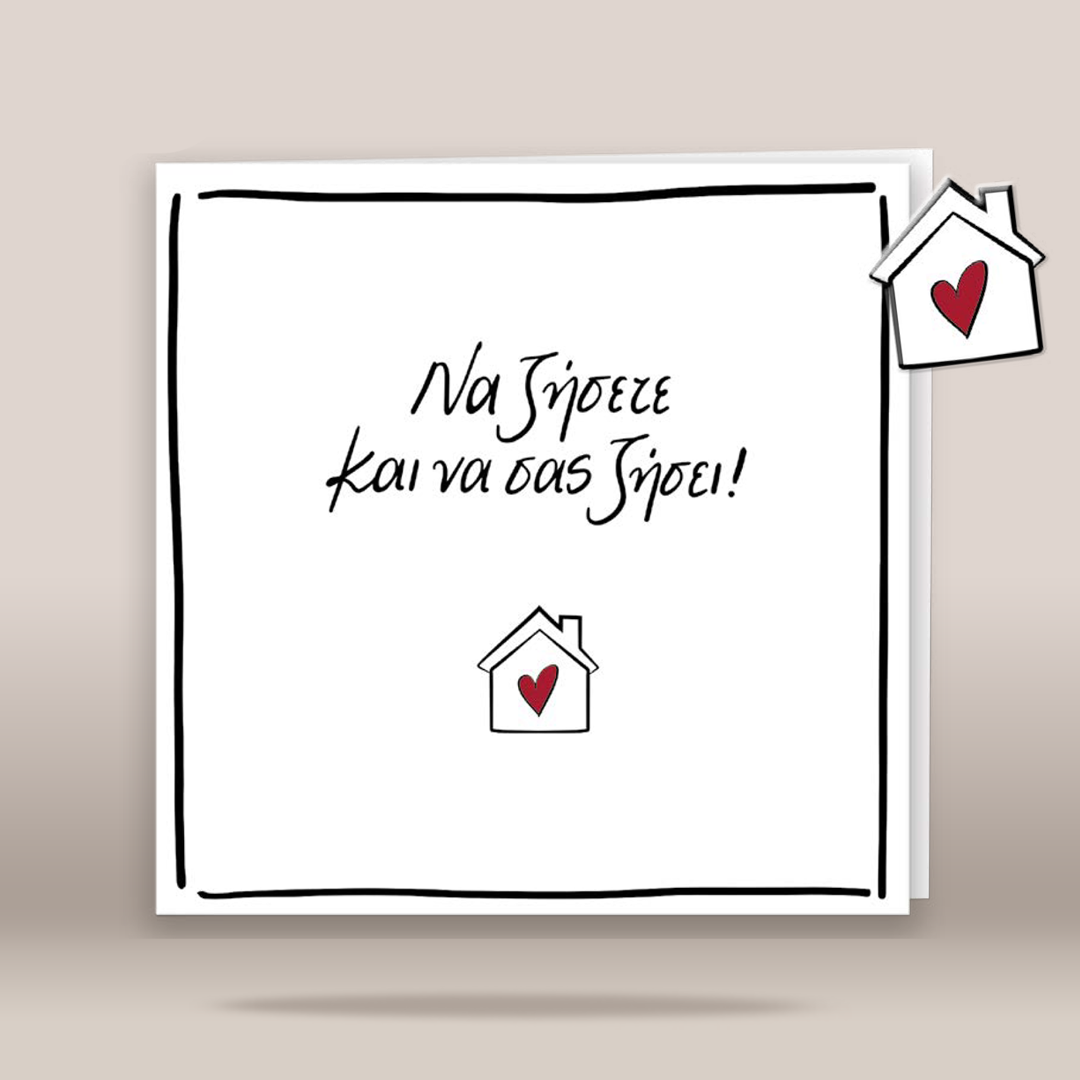 A square white greeting card with a hand-drawn black border and Greek text reading 'Να ζήσετε και να σας ζήσει' in black script, accented with small red hearts in two house icons. The card is shown standing against a warm beige background.