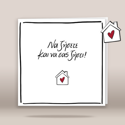 A square white greeting card with a hand-drawn black border and Greek text reading 'Να ζήσετε και να σας ζήσει' in black script, accented with small red hearts in two house icons. The card is shown standing against a warm beige background.