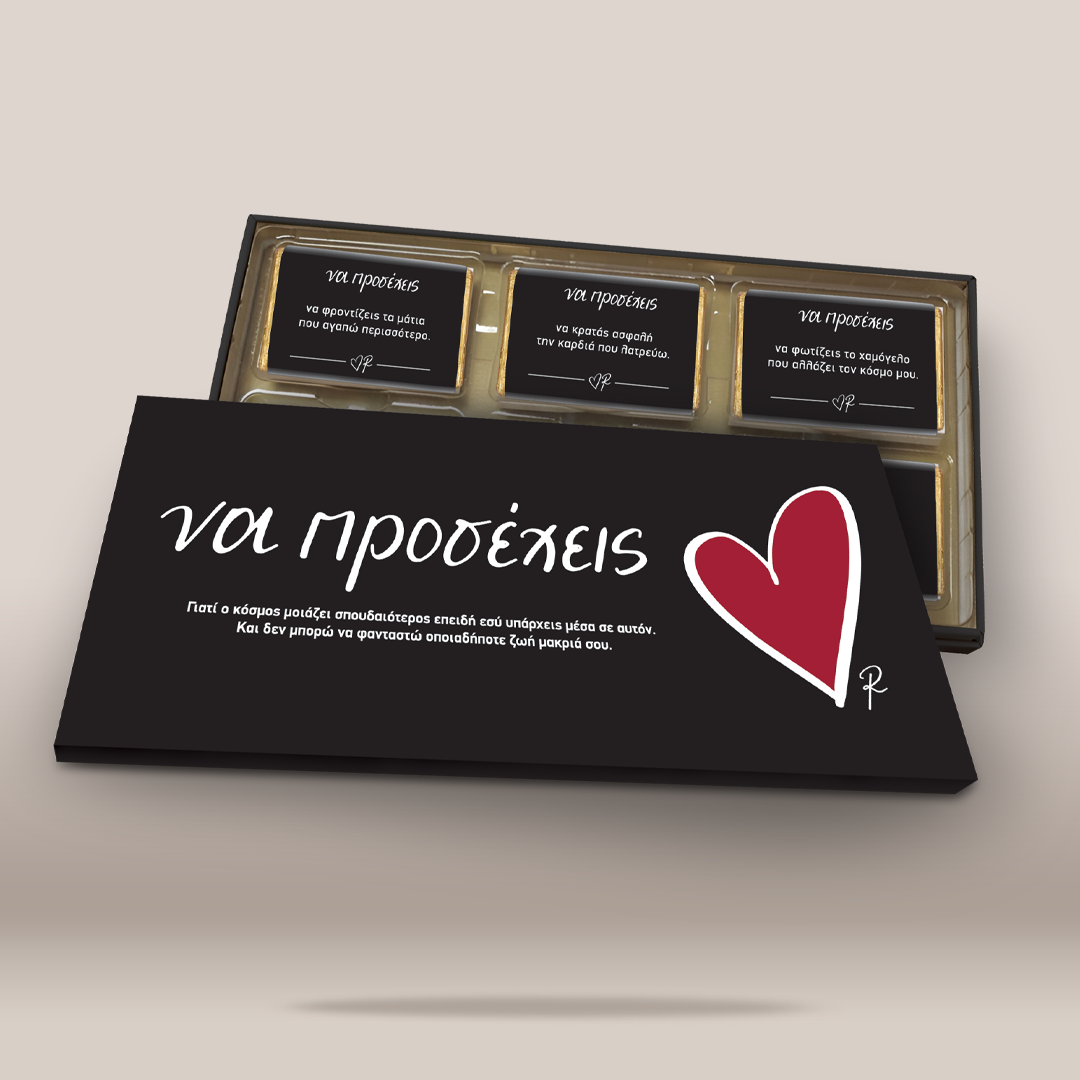 A black rectangular card or box cover with white Greek text and a red heart icon, placed in front of an open gold-trimmed case containing three small black cards with text, on a light beige background.