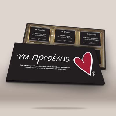 A black rectangular card or box cover with white Greek text and a red heart icon, placed in front of an open gold-trimmed case containing three small black cards with text, on a light beige background.