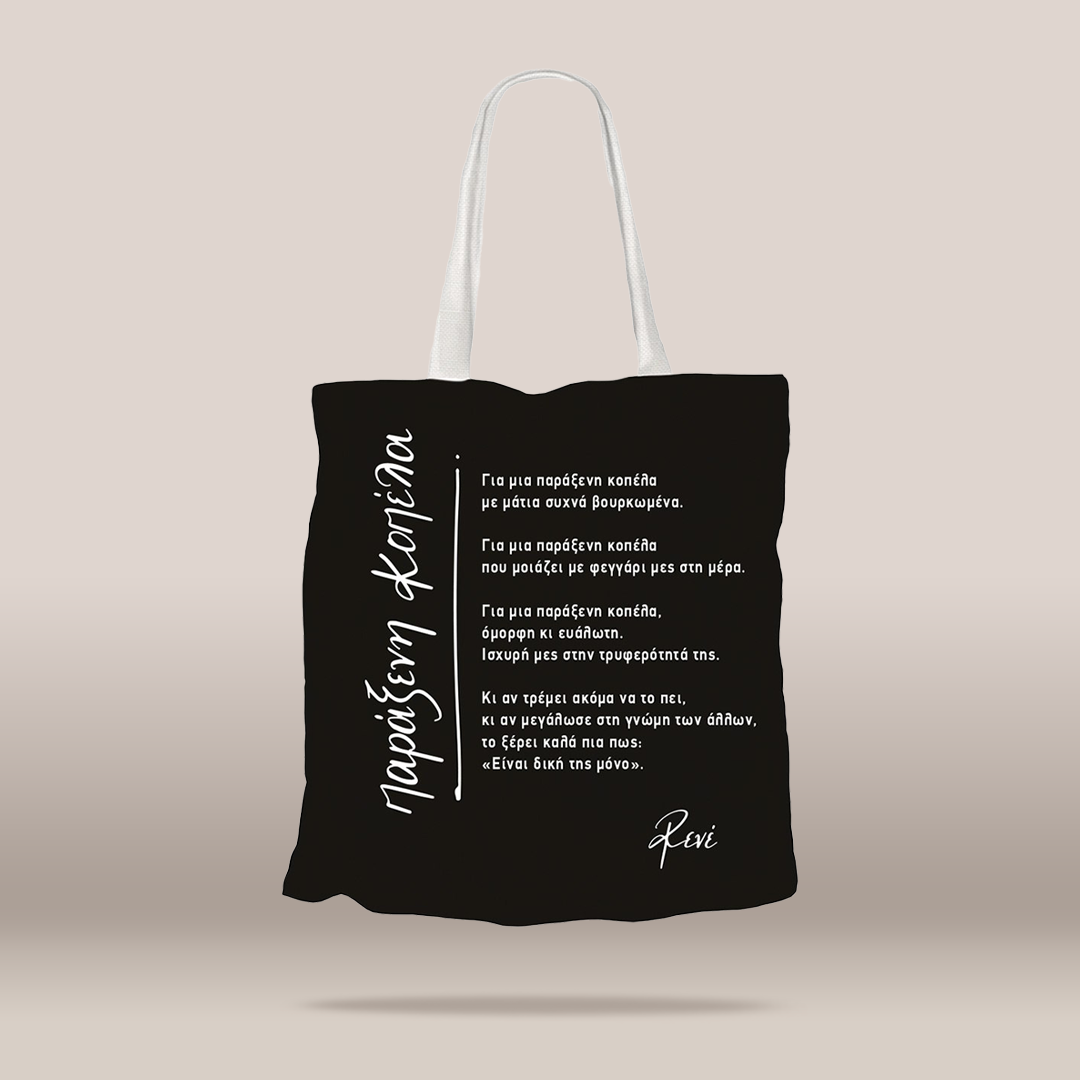 A black fabric tote bag with white handles, featuring white Greek text printed on the front, displayed against a neutral beige background.