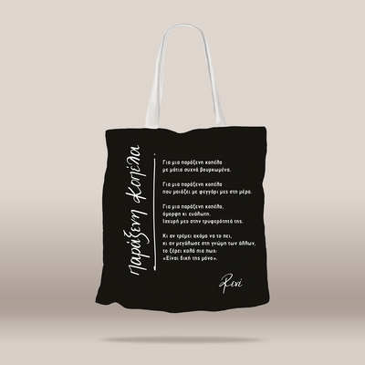 A black fabric tote bag with white handles, featuring white Greek text printed on the front, displayed against a neutral beige background.