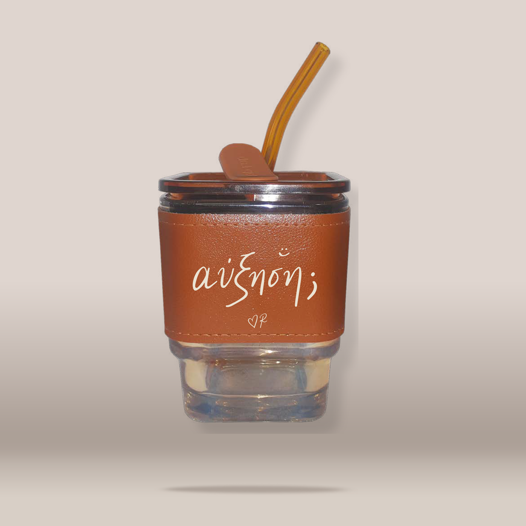 A clear glass tumbler with a brown faux-leather sleeve featuring the white script text 'ΑΥΞΗΣΗ', topped with a brown lid and a matching brown silicone straw, shown against a plain beige background.
