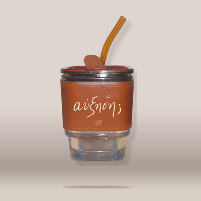 A clear glass tumbler with a brown faux-leather sleeve featuring the white script text 'ΑΥΞΗΣΗ', topped with a brown lid and a matching brown silicone straw, shown against a plain beige background.