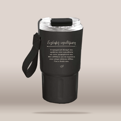 A matte black insulated travel tumbler with a clear lid, black carrying strap, and white Greek text printed on the front, shown on a plain beige background.