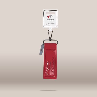 A red fabric keychain or bag charm with white printed text, attached to a silver metal clasp and hanging from a white branded card. 