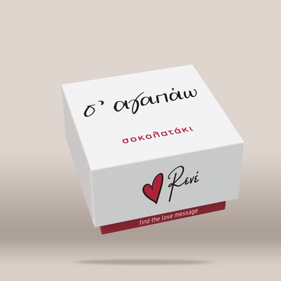 A white square gift box with Greek text on top and the Revé logo with a red heart on the front, shown floating against a warm beige background.