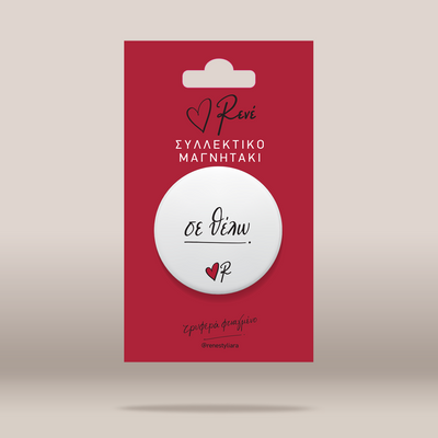 A white circular button badge with black script text and a small red heart logo, mounted on a red backing card with Greek text, shown on a light beige background.