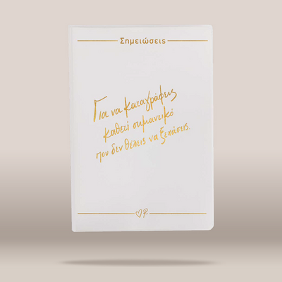 A white rectangular card or notebook cover with gold foil Greek text and thin gold lines, centered on a warm beige background.
