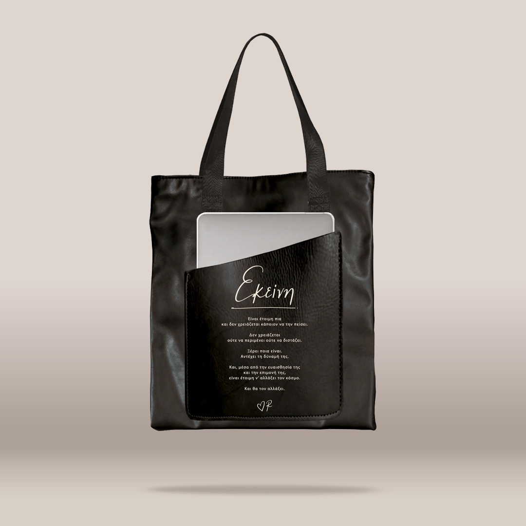A black faux-leather tote bag with a front pocket featuring a custom printed black card with white script text reading  and additional text below, shown against a solid beige background.