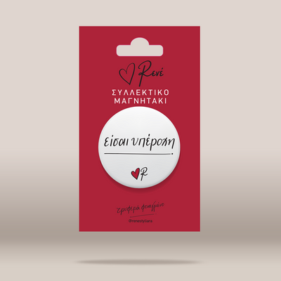 A white circular button badge with black cursive text and a small red heart logo, mounted on a red retail display card with Greek text, shown against a light beige background.